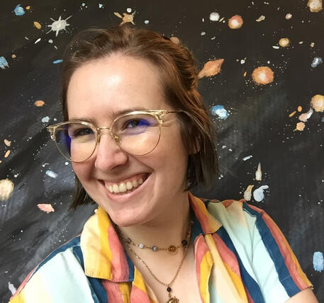 A picture of a person with white skin and cheek-length brown hair. They are wearing glasses, smiling wide, and wearing a striped shirt. They are standing in front of a space-themed background.