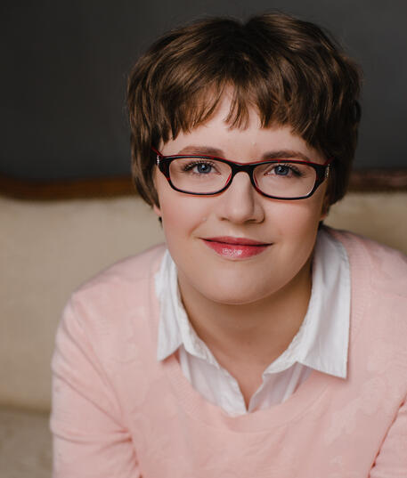 A picture of woman with short brown hair, white skin, glasses, and a light pink shirt.