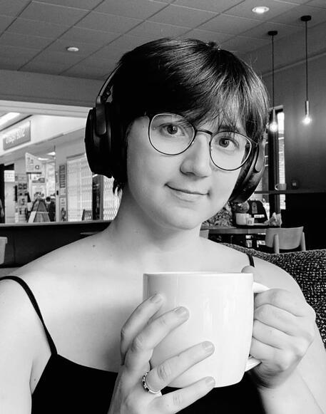 A grayscale picture of a person with white skin. They have short hair, headphones, and glasses. She is sitting with a mug in her hands, and is wearing a black tank top.